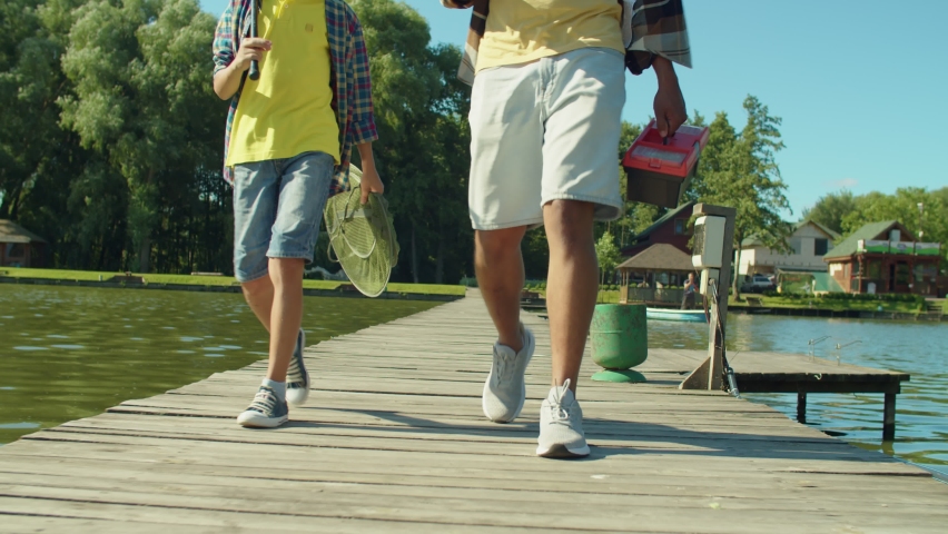 Low section of multiracial family with preadolescent son holding fishing rods, tackle box and equipment stepping on wooden pier, going for fishing on pond on summer day.