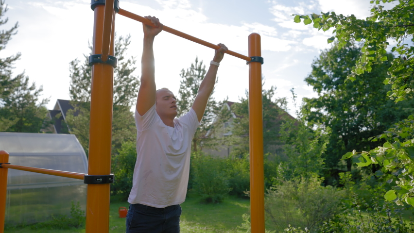 Mature man wearing white t-shirt catches up on yellow crossbar at back sunlight. Middle-aged sportsman enjoys doing sports in cottage house backyard