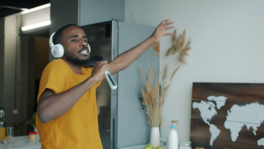 Playful young man singing in imaginary microphone listening to music through headphones and playing unreal guitar in kitchen at home.