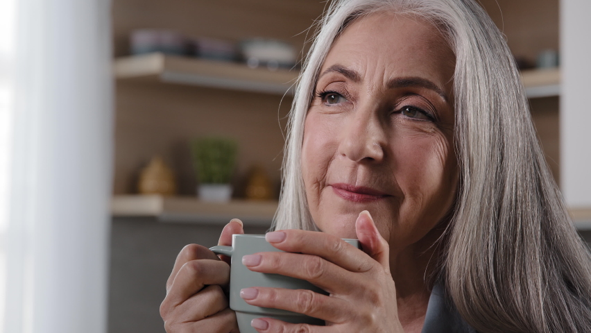 Dreamy inspired old blonde grey-haired woman granny grandmother mature middle aged woman pensioner sitting in kitchen drinking coffee tea hot drink healthy liquid from cup daydreaming relaxing at | Shutterstock HD Video #1093217357