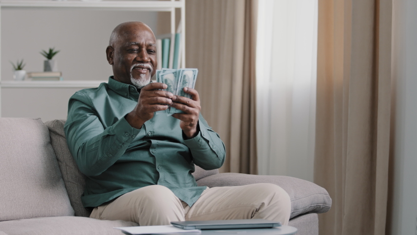Happy old bald overweight African American senior mature elderly man happy with financial savings salary at home sitting couch. Winning online lottery holding dollars throw up cash money rain Royalty-Free Stock Footage #1093217367