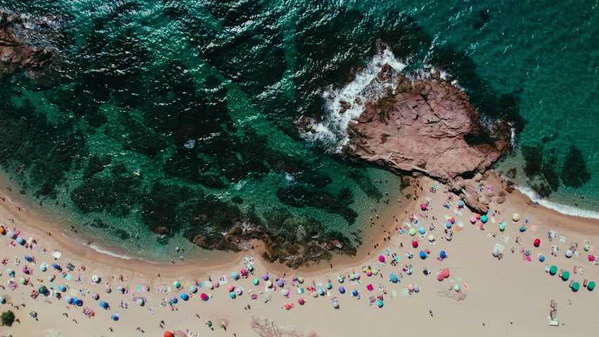 Top birds eye view on secluded beautiful cove in mediterranean sea on spanish coast. Gentle waves crash on rocks and sandy beach. Holiday goers on vacation under sun umbrellas relax and tan