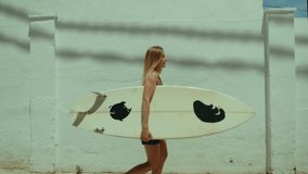 Side tracking shot of young blonde woman walk towards beach with surfboard. Surfing town summertime vacation. Female surfer beach lifestyle. Woman surfer enjoy summer breeze in portugal  - Powered by Shutterstock - Get 15% off with code: PIKWIZARD15