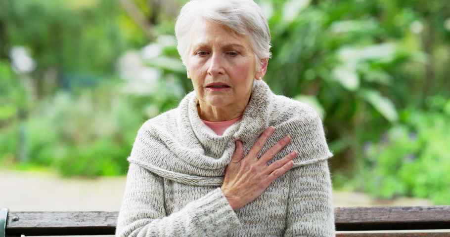 Senior woman suffering from heart attack, breathing heavily and holding her chest in pain. Mature female experiencing a cardiac arrest and looking scared or afraid while sitting on a bench on a park | Shutterstock HD Video #1093218165