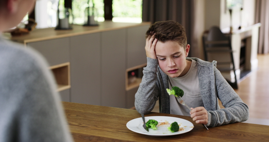 Frustrated boy told to eat healthy vegetables that he hates eating by his mom at meal time on his dinner plate. Mother stands in the kitchen to watch her unhappy and upset child finish his broccoli. | Shutterstock HD Video #1093218197