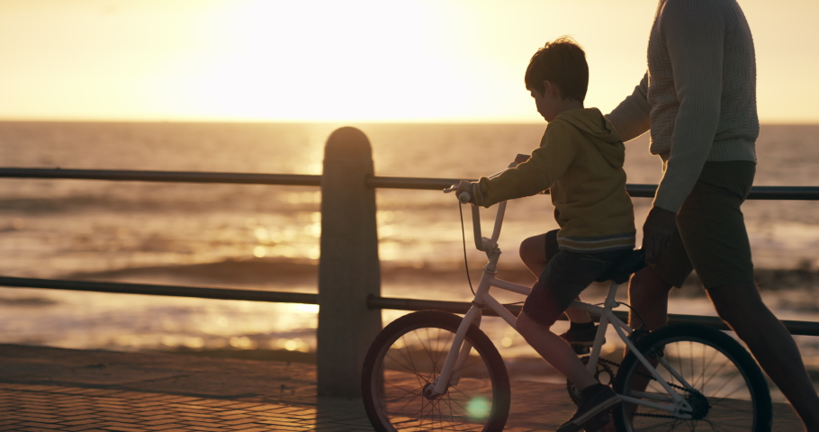 Learning to ride a bike with a father teaching his son to cycle at the beach by the sea. Young boy child on his bicycle with his dad, having fun and bonding together as a family at sunset in summer Royalty-Free Stock Footage #1093218255