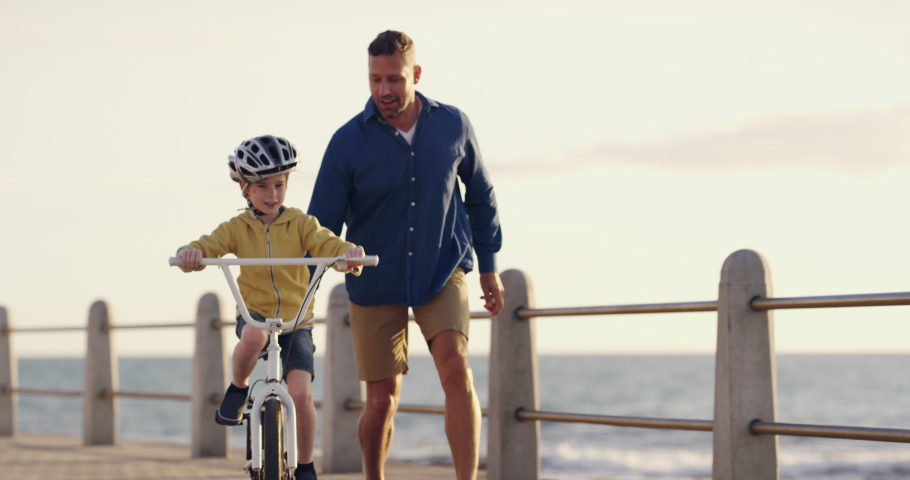 Learning, bicycle and proud dad teaching his young son to ride while wearing a helmet for safety along a seaside promenade. Active father helping and supporting his child while cycling at sunset Royalty-Free Stock Footage #1093218293