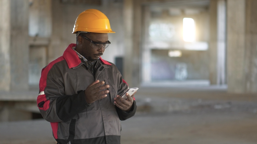 Slow Motion African american workman in yellow hard hat stands at construction site, holds in hands smartphone and dials number