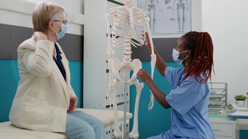 Osteopath nurse examining human skeleton bones with patient at consultation appointment, explaining orthopedic disease and diagnosis. Medical assistant and woman during coronavirus pandemic. | Shutterstock HD Video #1093219465