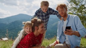 Joyful family using smartphone making photo in summer mountains close up. Happy couple with children waving hands to mobile phone camera sitting green grass. Cheerful parents have fun with kids.