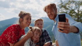 Happy family having video call on sunny nature. Smiling young parents with children using cellphone in summer mountains. Cheerful couple with kids speaking on mobile phone sitting beautiful meadow.