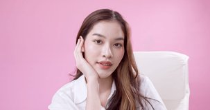 Video of an Asian girl who is shy but happy sits on a white chair against a pink background and exhibits happiness while wearing white clothing.