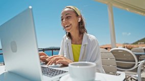 Happy woman in earphones laughing and speaking with someone by laptop outdoors on summer terrace with beautiful sea view, video call with family from hotel on resort during travel