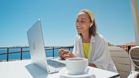 Happy woman in earphones laughing and speaking with someone by laptop outdoors on summer terrace with beautiful sea view, video call with family from hotel on resort during travel - Powered by Shutterstock - Get 15% off with code: PIKWIZARD15