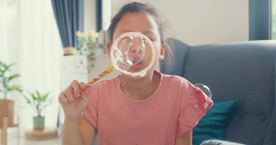 Happy Asian toddler little girl sit on sofa chair play blowing bubbles in front of camera with natural smile having fun and joyful in living room at home. Creative lifestyle for kid concept.
