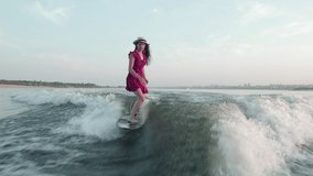A female surfer in a red dress and hat jumps on a wakeboard. An experienced wakeboarder sprays water drops into the camera.