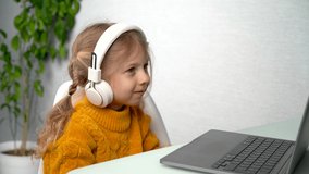 online learning, a little blonde girl in white headphones learns at a laptop, listens to music and dances