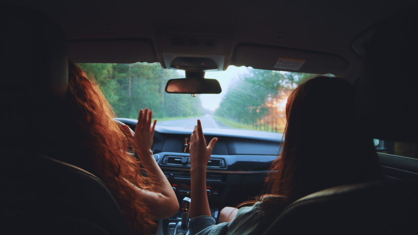 Two happy female woman friends enjoy travel in car. Sitting in front seat and have fun music song on road trip. Girls driving car and dancing. Concept of youth, friendship, holidays, sunset vacation.