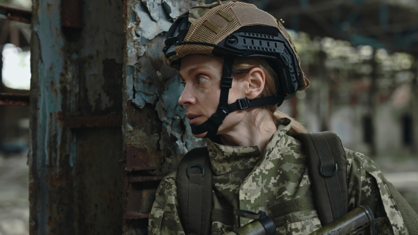Caucasian woman in military uniform standing on damaged manufacture plant with fear on face during enemy occupation. Female soldier with weapon hiding behind column.
