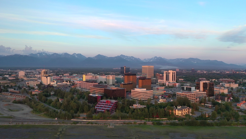 Aerial drone view of Anchorage Alaska skyline at sunset in summer. Beautiful birds eye footage captures the glowing cityscape with high rise buildings downtown core and dramatic orange pink sunset light over the urban landscape with mountain backdrop. Shot in crisp 4K this dynamic clip showcases the vibrant evening atmosphere of Alaskas largest city. Perfect for travel documentaries Alaska tourism promotions cityscape footage sunset aerials urban lifestyle content or stock videos about Anchorage and northern summer evenings.