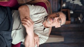 VERTICAL VIDEO POV portrait confident gray haired grandfather posing in red vintage armchair with crossed hands. Focused elderly aged man in eyeglasses relaxing at rustic house enjoy retirement