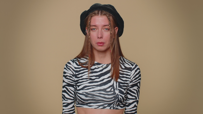 Pretty young woman pointing around with finger gun gesture, looking confident, making choice, shooting killing with hand pistol right on target. Adult stylish girl isolated on beige studio background