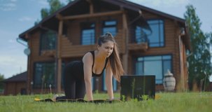 Young athletic girl performs leg training exercises on fitness mat using laptop. Online training in fresh air. Slow motion