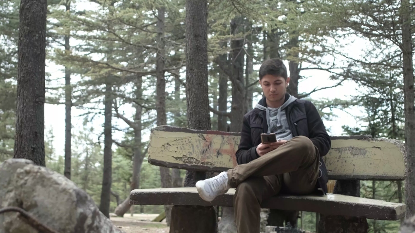 Young man sitting and relaxing on green forest bench, view of man spending time listening to music in trees, enjoying nature by listening to music with headphones
