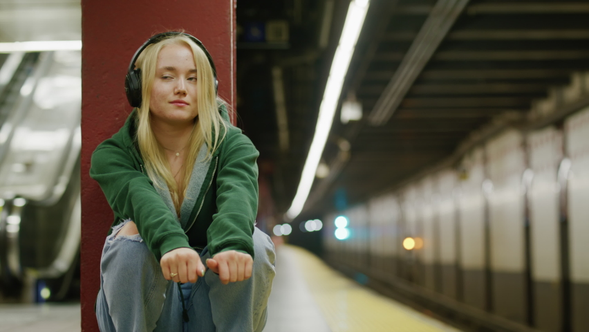 Female commuter waiting for a train at the NYC subway station with escalator in the background. Cheerful, blonde-haired woman in headphones in green casual jacket underground. High quality 4k Royalty-Free Stock Footage #1093294341