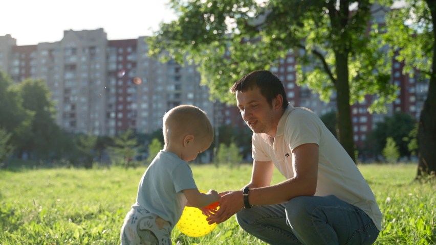 Toddler son and father play with bright balls in green city park. Father wearing white t-shirt and blue jeans shows and touches red ball to child closeup