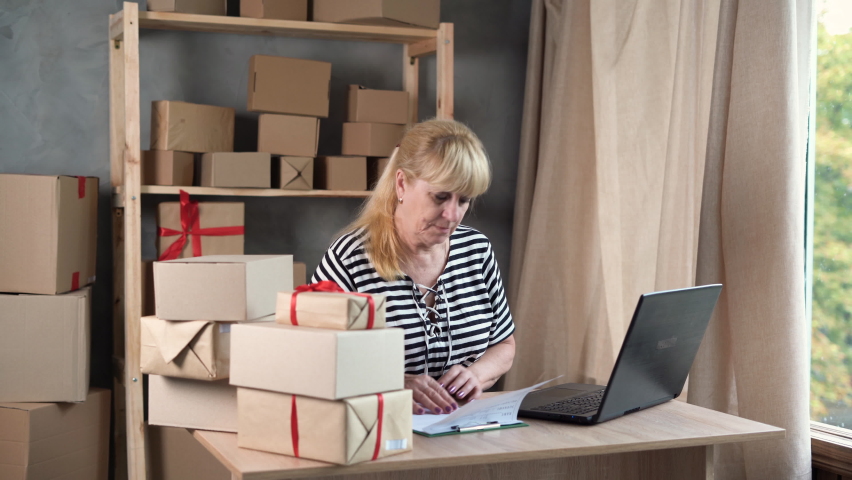 Older female entrepreneur online store owner using laptop at work preparing parcel boxes, checking post shipping online retail e-commerce store order in dropshipping delivery warehouse. SME Royalty-Free Stock Footage #1093309451