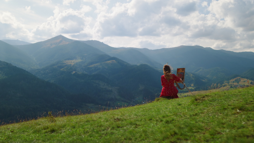 Back view of woman painter painting picture of gorgeous mountain landscape outdoors. Unknown girl drawing on easel sitting green hill summer holiday. Unrecognizable talented artist working on nature