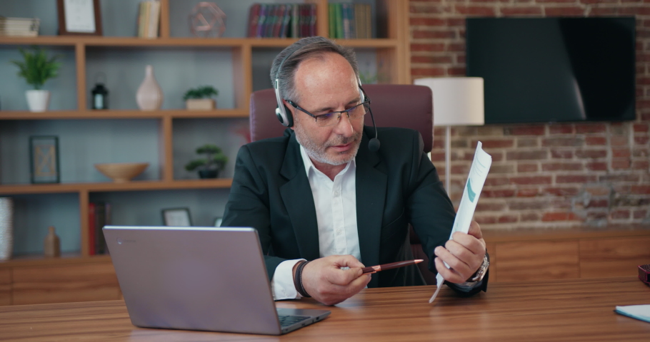 Handsome experienced purposeful adult bearded businessman in headset holding a videochat with colleagues on laptop and explaining financial report Royalty-Free Stock Footage #1093312731