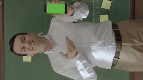 Male teacher looking at information on phone in front of blackboard, searching for something online, reading news, checking social networks.The teacher shows his green screen smartphone to the camera.