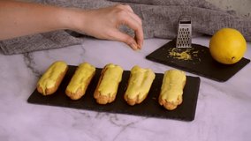 Eclairs with custard and lemon glaze. A woman's hand sprinkles eclairs with lemon zest. Confectionery for sweet tooth. The concept of confectionery video.