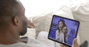 Young and happy african man sitting on sofa in living room at home use tablet making video call for talking with his girlfriend and waving hand to dog while city lockdown. Long distance relationship.