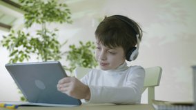 Little Smiling Boy Sitting At The Table Listening To Music And Playing Games On The Digital Tablet Watching Video Surfing Internet