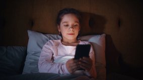 Small Girl Using Smartphone in Bed Before Going to Sleep. Cute Kid Checking Social Media, School Schedule, Reading Homework on Digital Ebook, Chatting with Friends Online.