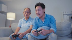 Asian happy family play console game together in living room at night. Senior elderly older father and young handsome male sit on sofa, feel excited and enjoy spend time play joystick gaming in house.