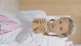 Vertical video. Cute little girl holding cardboard cup and drinking cacao or hot chocolate from a straw indoors