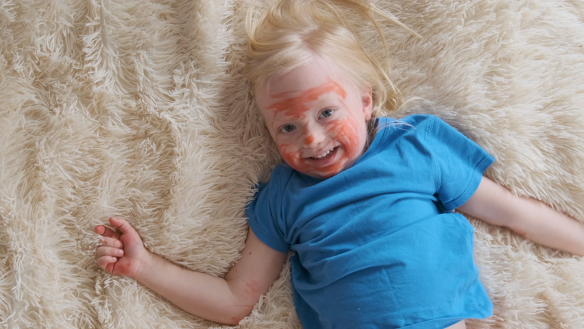 Little girl lies on soft blanket and makes faces. Blonde child with red face and hands from felt-tip pen.