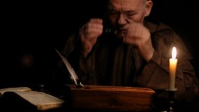 Video of old monk takes a manuscript out of the document box, signs it, wet paperweight, and puts it back. Priest in glasses and monk robe in dark monastic cell room under candle light. Zoom effect