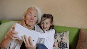 Loving elderly grandmother and granddaughter on the sofa, in the living room using a tablet together. A smiling granny and a girl have fun with a notebook. The concept of digital technology.