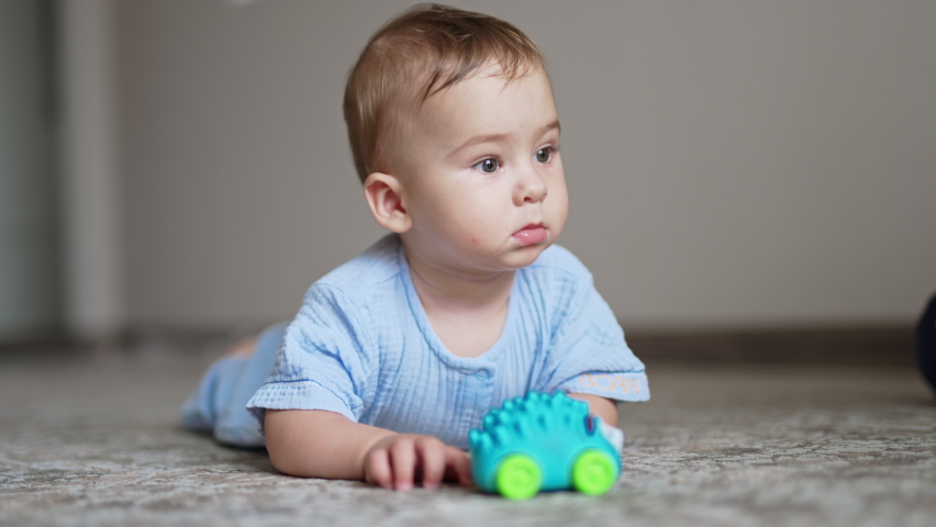 Beautiful baby lying on belly on the floor with toy in front of him. Cute child looks up and smiles to somebody adorably. Blurred backdrop. | Shutterstock HD Video #1093368895