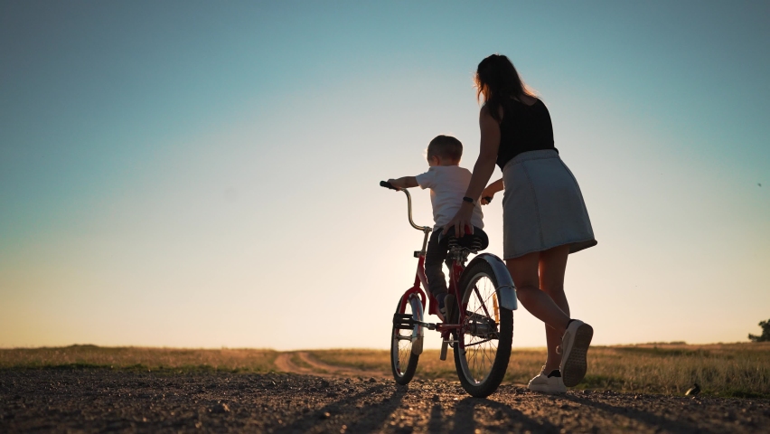 Happy family concept. Mom teaches her son to ride bike on green grass. Child rides bicycle along rural road. Green energy. Mom teaches her son to ride bike for the first time in park at sunset. Royalty-Free Stock Footage #1093369713