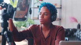 Young energetic African American woman blogger conducts video broadcast recording himself on phone camera communicates emotionally with channel subscribers sits at table with microphone in home office