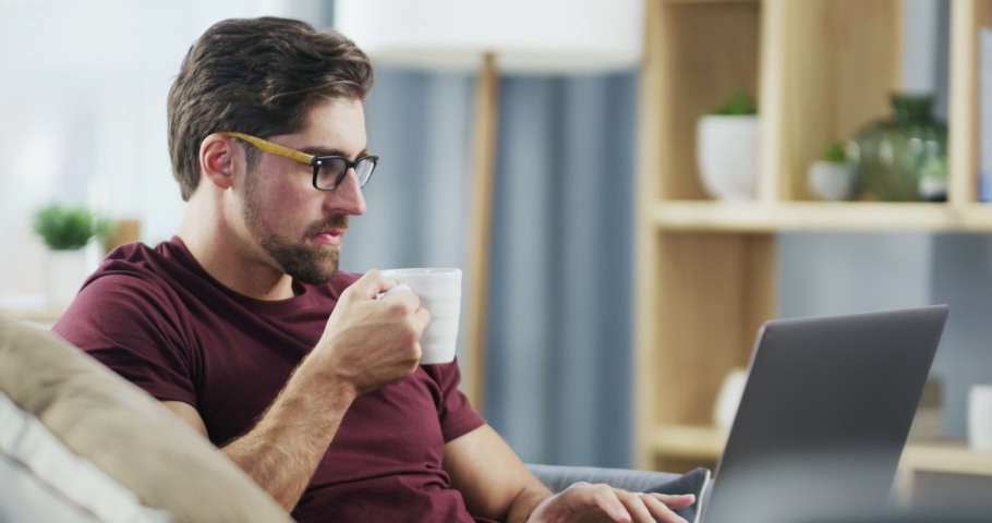 Male entrepreneur working online on a computer while drinking coffee sitting on a couch at home. Remote digital freelance worker typing an email and doing web research on a laptop indoors | Shutterstock HD Video #1093375203