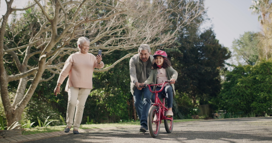 Excited, happy little girl learning to ride a bicycle with her excited grandparents. Proud grandmother using a phone to take a picture of a special moment while bonding with grandchild outdoors | Shutterstock HD Video #1093375251