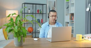 Hardworking asian man wear wireless headset making conference video call on laptop. Male professional call center agent, hr manager having distance webcam chat job interview on computer in home