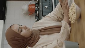 Young woman in hijab works remotely on a tablet computer. The woman drinks coffee or tea and eats roasted hazelnuts while working on the computer in her home kitchen. Freelancer concept.Vertical video
